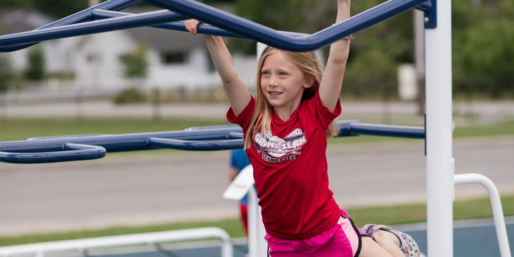 Child using monkey bars at outdoor playground fitness structure designed for upper body strength and coordination